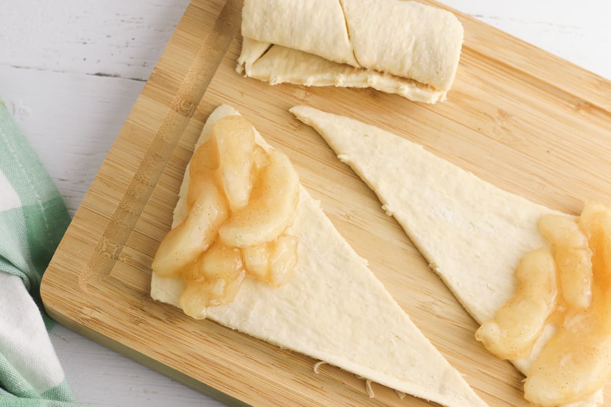 Two triangles of raw crescent roll dough on a cutting board. A spoonful of apple pie filling in on the wide end of each 