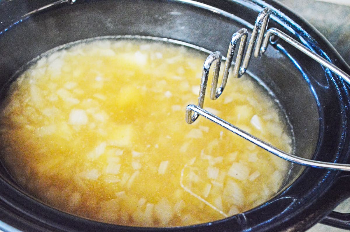 potato masher over crockpot filled with chicken broth, salt, pepper, onion, and mashed potatoes.