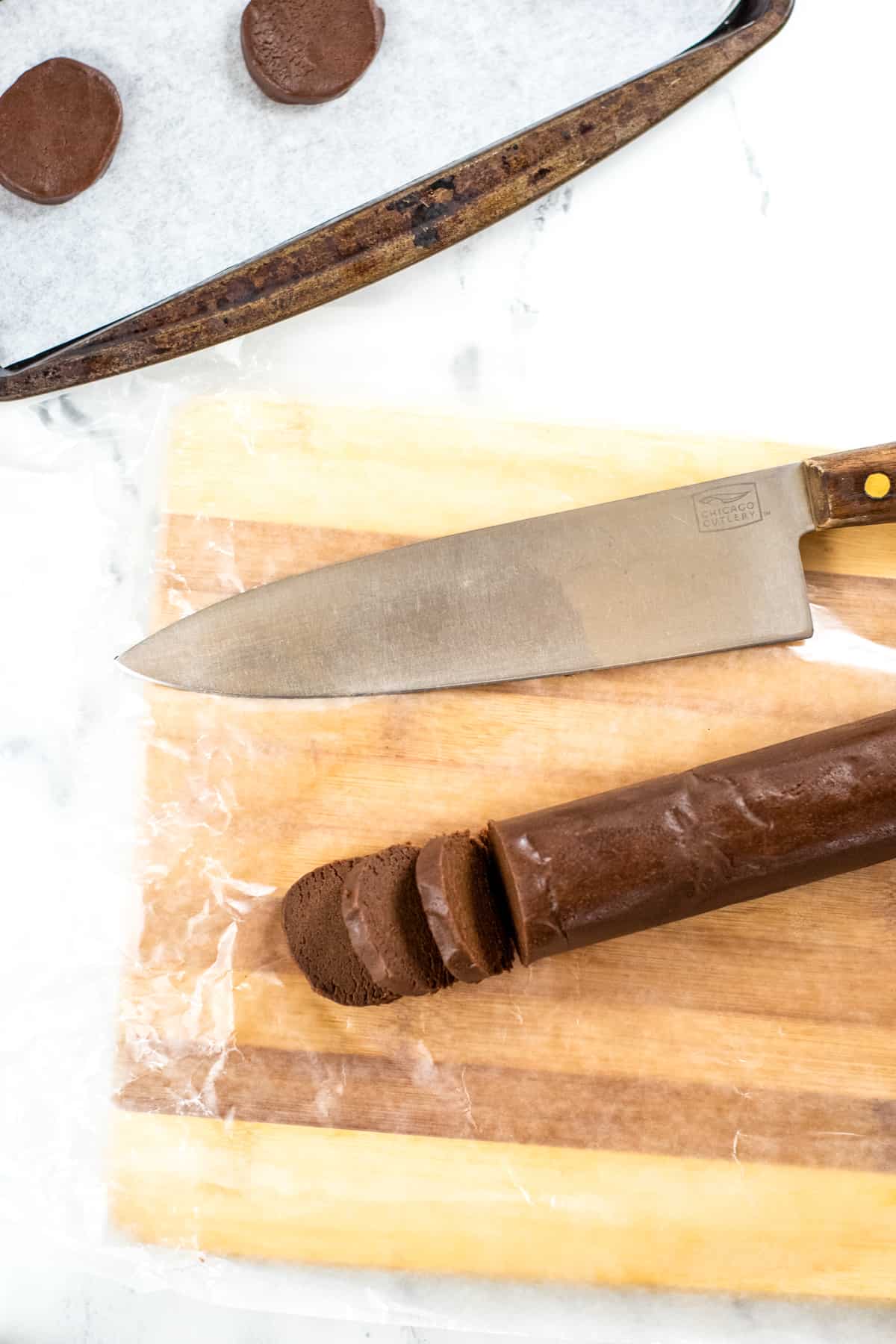 Cutting board with log of chocolate cookie dough, some of which as been cut into slices. Large knife is on cutting board and lined baking sheet with slices of cookies are shown as well.