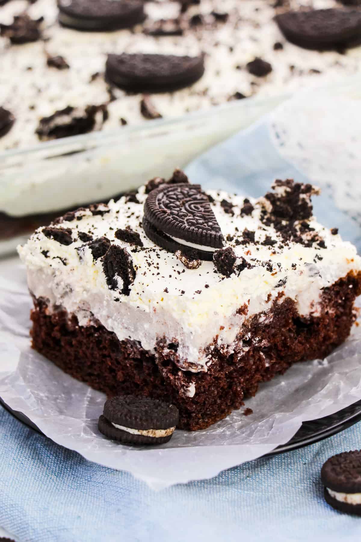 Square slice of OREO Poke Cake topped with Cool Whip and pieces of OREO cookies. Whole OREO cake in 9 x 13 baking dish behind plate.