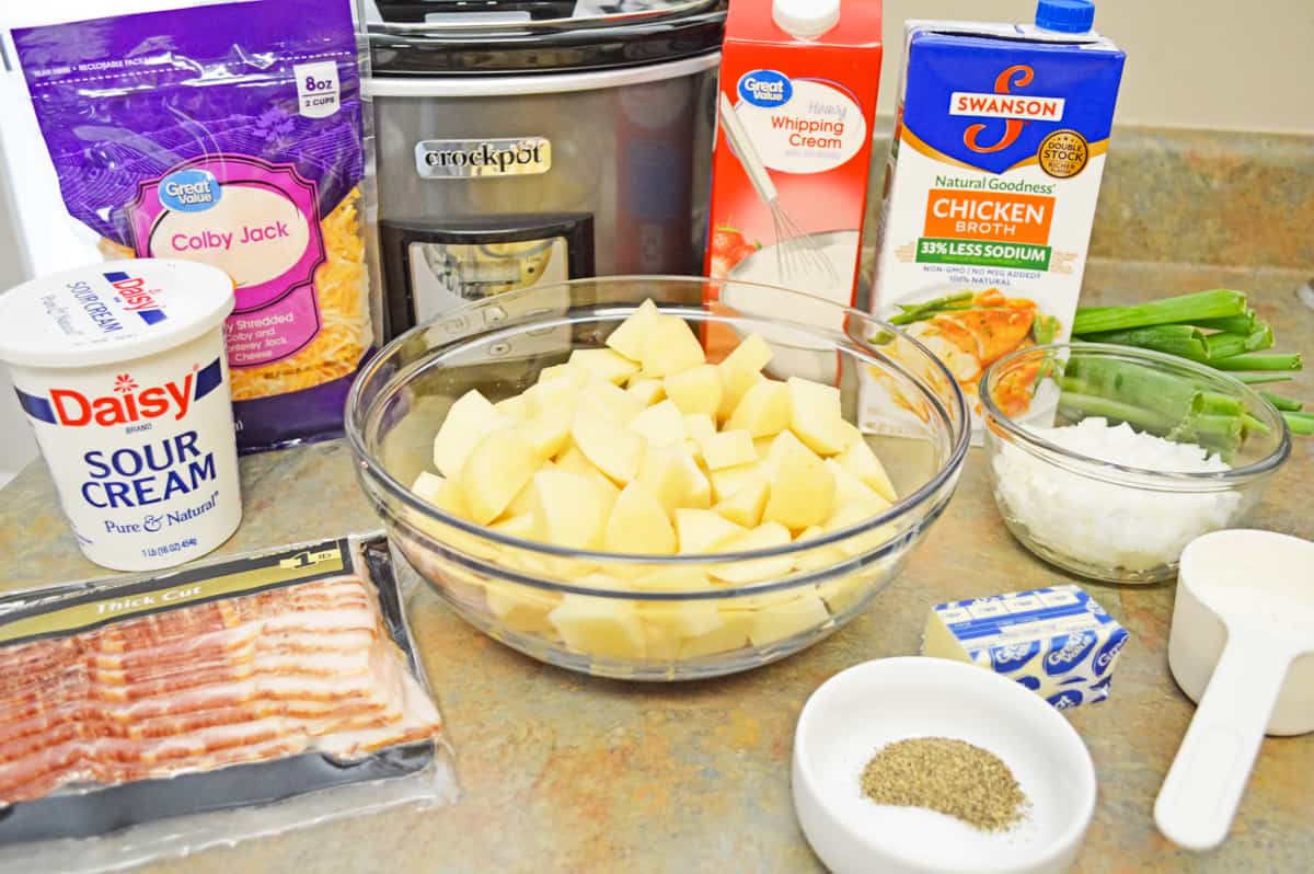 Ingredients on countertop: shredded cheese, sour cream, bacon, chopped potatoes, heavy cream, chicken broth, chopped onion, green onions, salt, pepper, butter, and flour