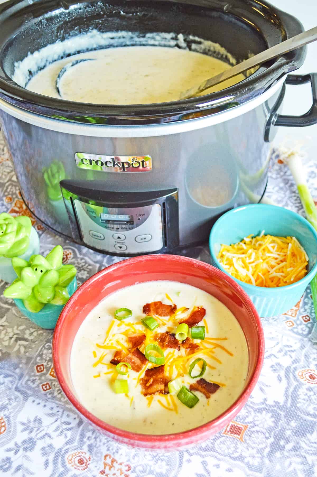 Crockpot full of baked potato soup with bowl of soup topped with scallions, cheese, and bacon next to it. To the side are a bowl of shredded cheese and salt and pepper shakers
