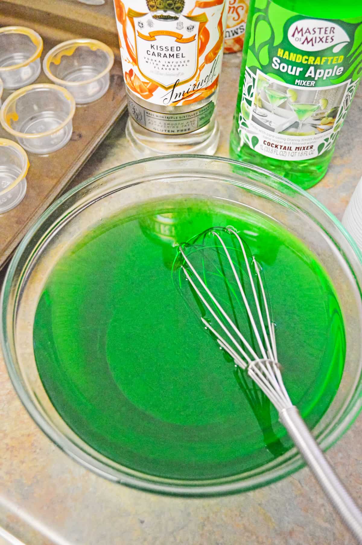 Glass bowl with green jello and wire whisk. Empty shot glasses, bottle of caramel vodka, and sour apple mixer are in background