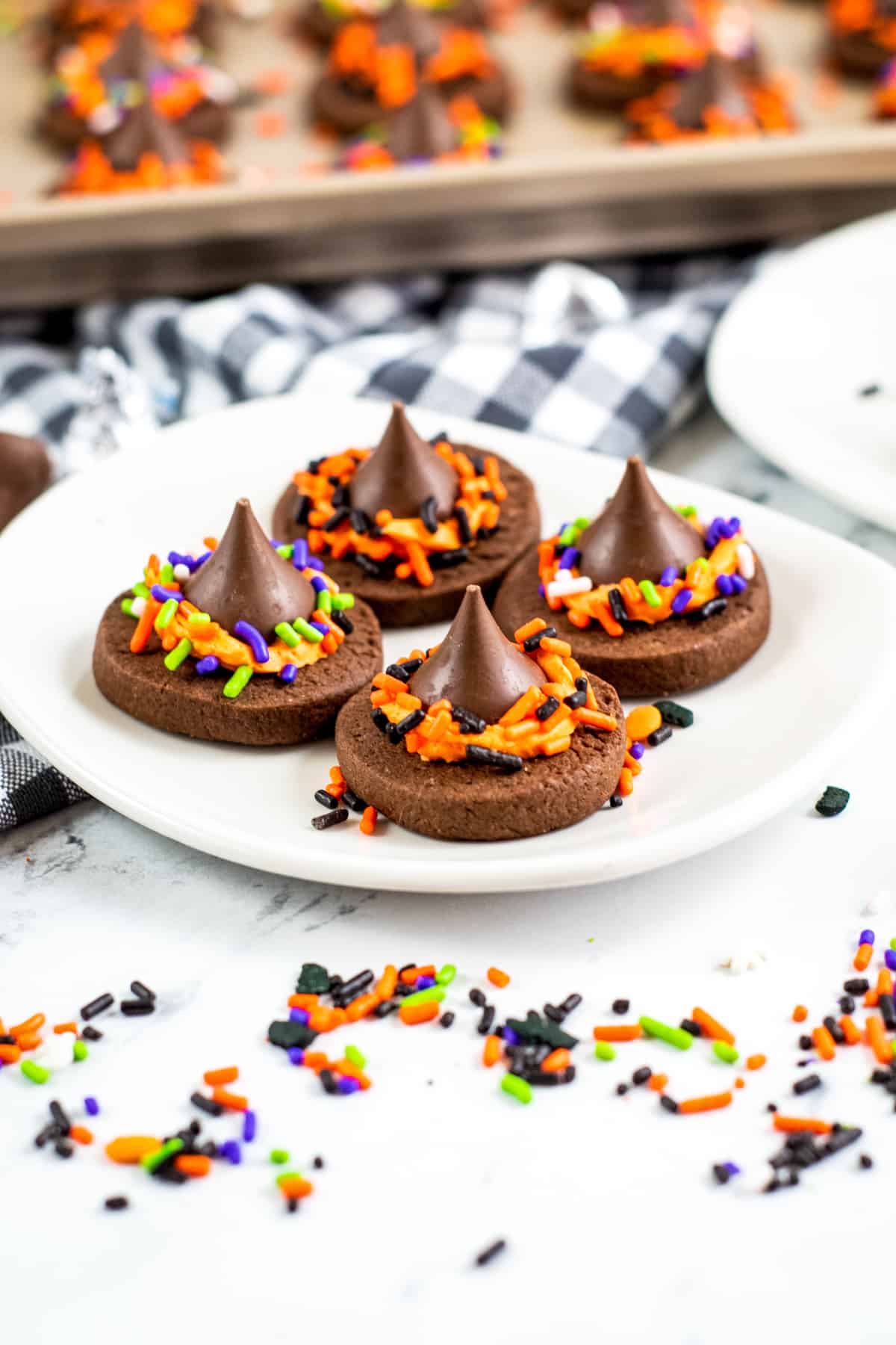 Four chocolate witch hat cookies with Hershey kisses on a white plate with halloween sprinkles around the table top and more witch hat cookies in the background.