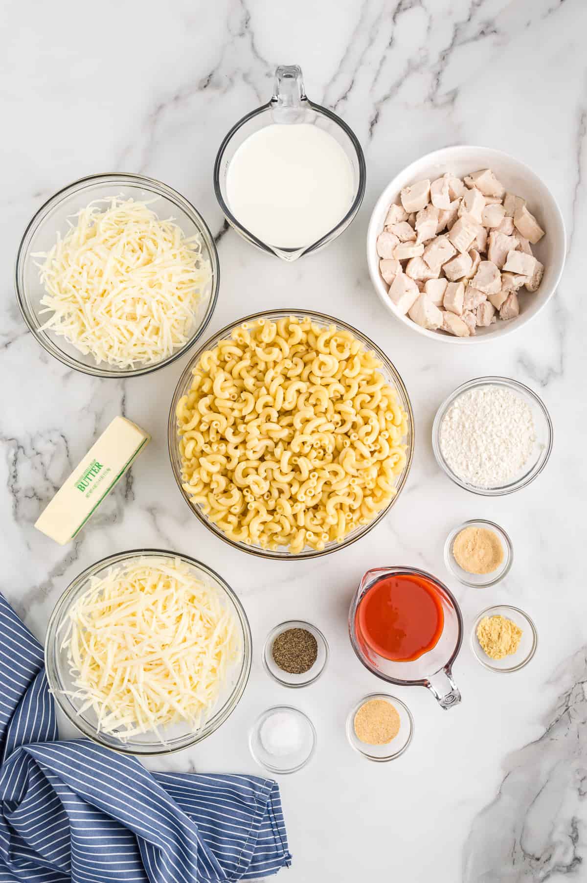 Ingredients on marble countertop: bowl of shredded cheese, measuring cup of milk, bowl of cubed cooked chicken, bowl of flour, measuring cup of buffalo sauce, 4 small bowls of spices and seasonings, a second bowl of shredded cheese, one stick of butter, and bowl of elbow macaroni.