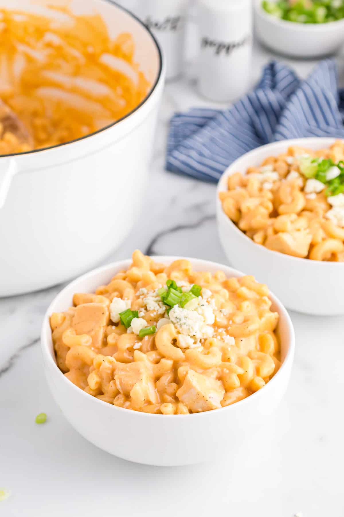 Buffalo Chicken Mac and Cheese in white bowl topped with blue cheese and scallions. Another serving and a large pot with the rest of the pasta can be seen in background.