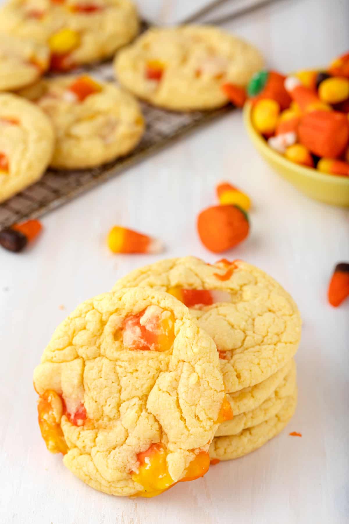 Candy Corn Cake Mix Cookies stacked on tabletop with one cookie on its side to show candy corn inside them. Extra candy corn and more cookies are on cooling rack in background.