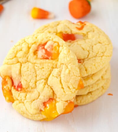 Candy CornCookies stacked on tabletop with one cookie on its side to show candy corn inside them. Extra candy corn are scattered in background.