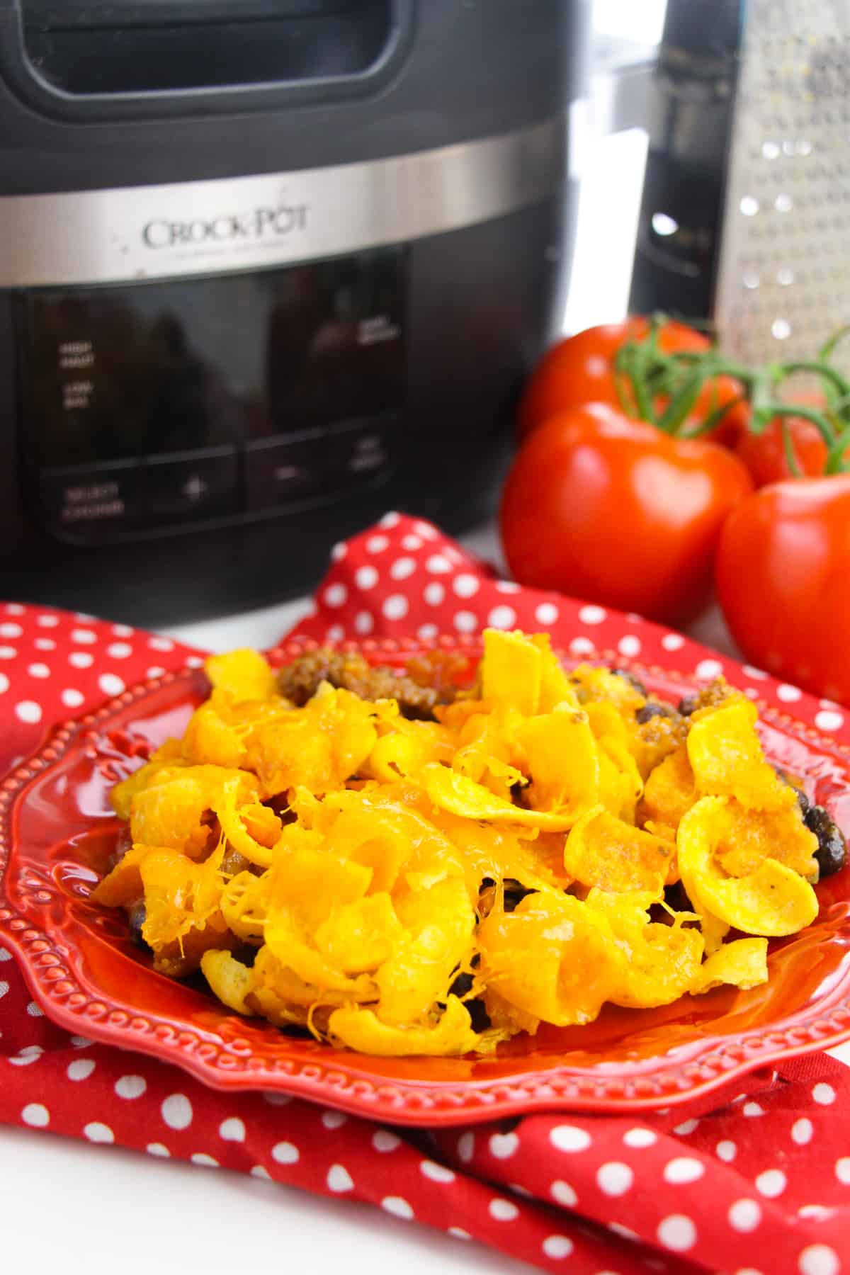 Crockpot Frito Pie served on red plate with slow cooker, tomatoes, and cheese grater in background