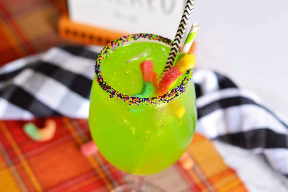 Bright green drink in Halloween sprinkle-rimmed wine glass with 2 gummy words and 2 paper straws sticking out of it.