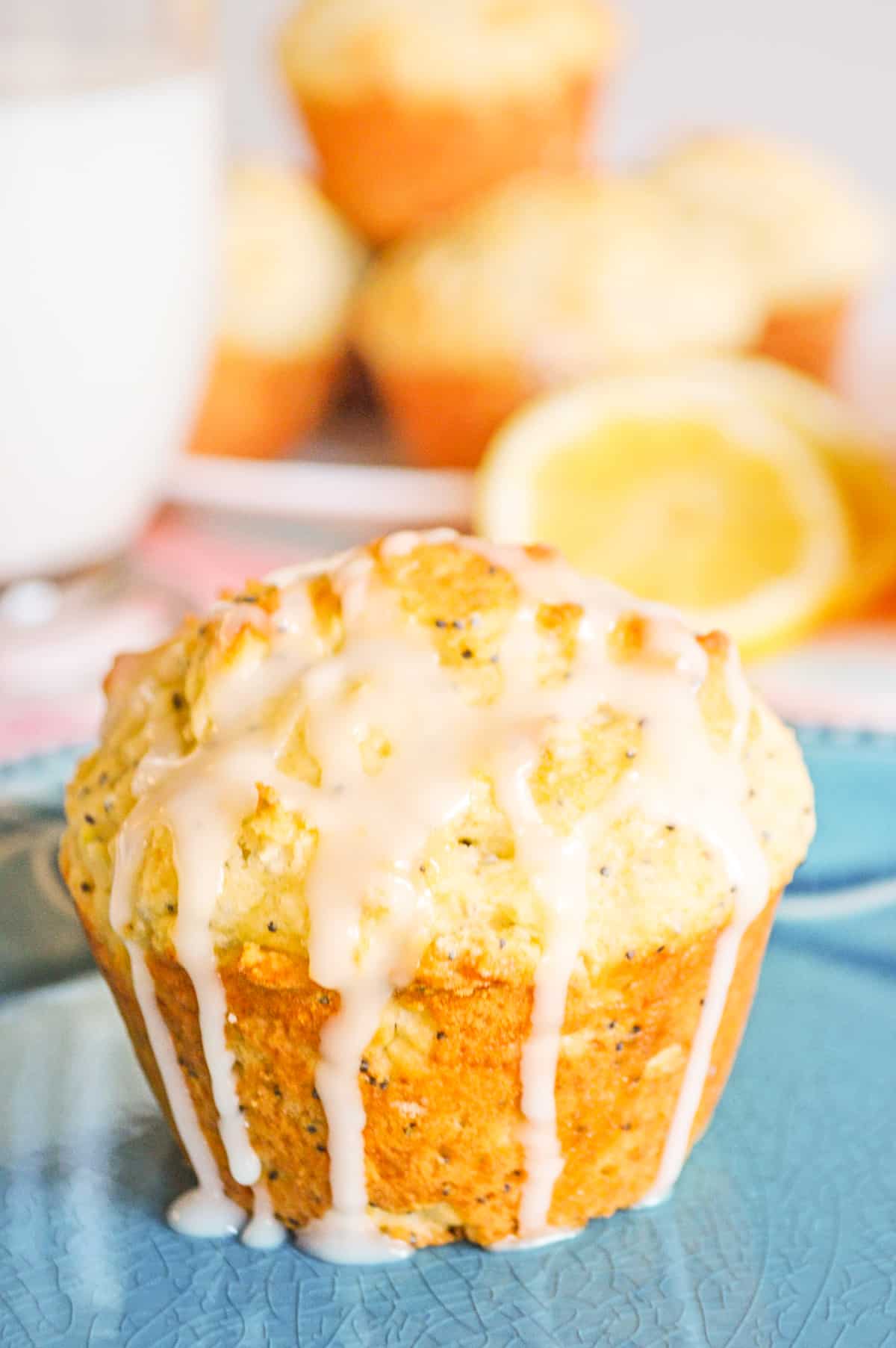 Lemon poppy seed muffin drizzled with lemon glaze which is dripping down the sides. Additional muffins and sliced lemons are behind the muffin.