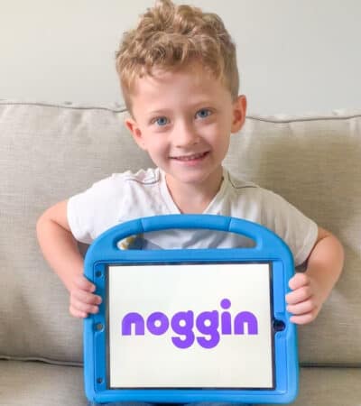 4-year-old boy sitting on a couch and holding up an ipad in a blue plastic case with ipad screen reading: noggin