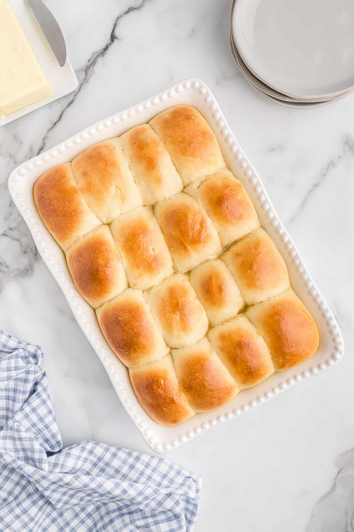 16 golden brown potato dinner rolls in white baking dish with plates, linen, and butter around them