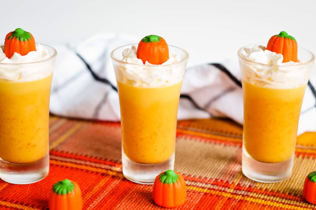 Pumpkin jello shots in glass shot glasses, topped with whipped cream and candy pumpkins. More candy pumpkins are scattered on tablecloth.