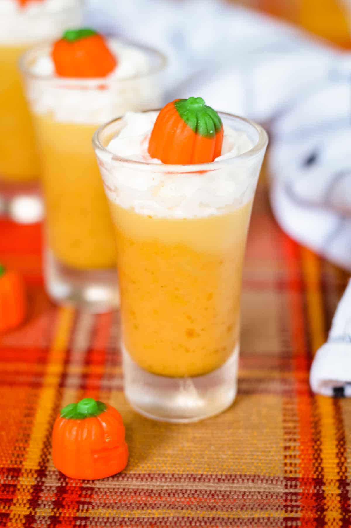 Pumpkin Pie Jello Shot in tall glass shot glass. The thick, orange shot is topped with whipped cream and a candy pumpkin.