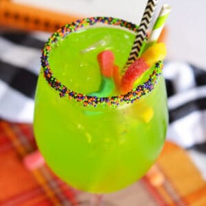 Bright green drink in Halloween sprinkle-rimmed wine glass with 2 gummy words and 2 paper straws sticking out of it.