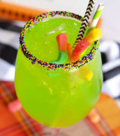 Bright green drink in Halloween sprinkle-rimmed wine glass with 2 gummy words and 2 paper straws sticking out of it.