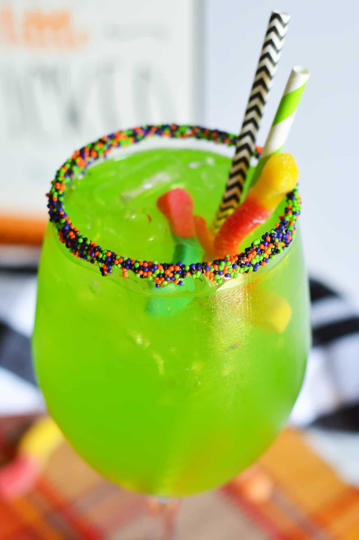 Witches Brew Cocktail- bright green drink with colorful gummy worms hanging out of it. Served in a wine glass with Halloween sprinkles on the rim and Halloween paper straws.