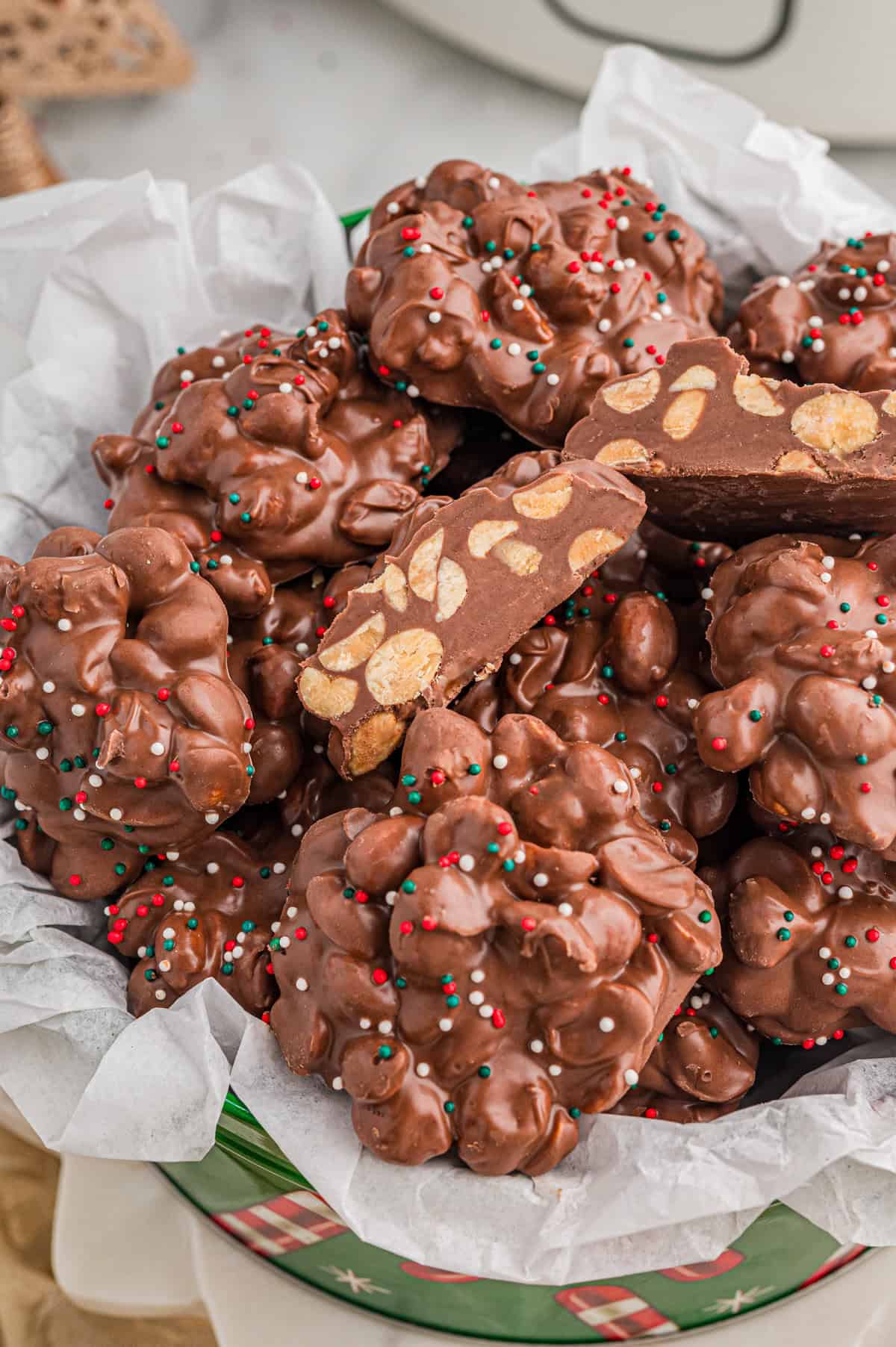 Christmas tin lined with parchment paper and filled with chocolate peanut clusters - "crockpot candy", each topped with sprinkles. One broken cluster reveals whole peanuts inside.
