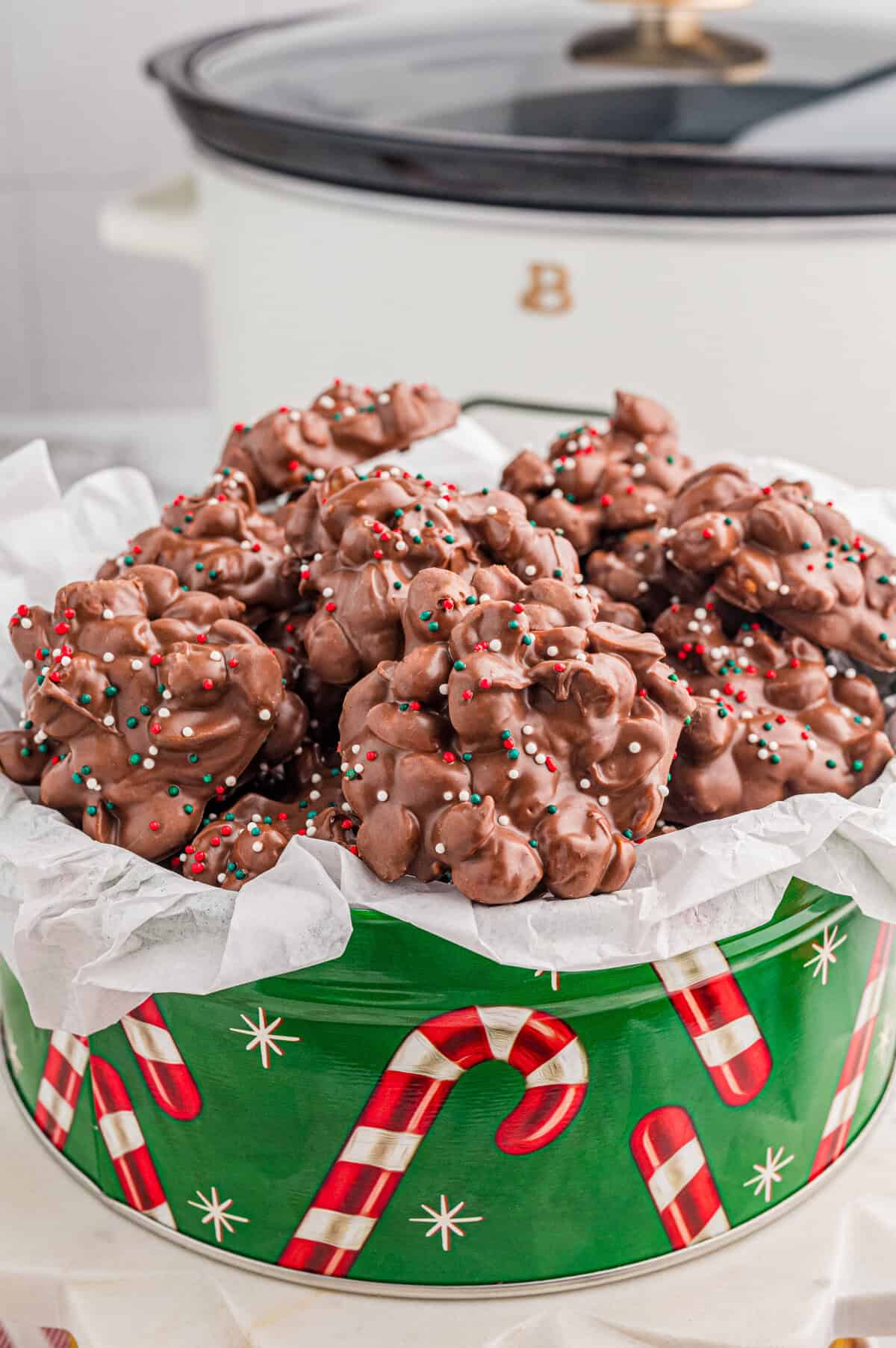 Crock pot Christmas candy in a festive green tin with slow cooker in the background.