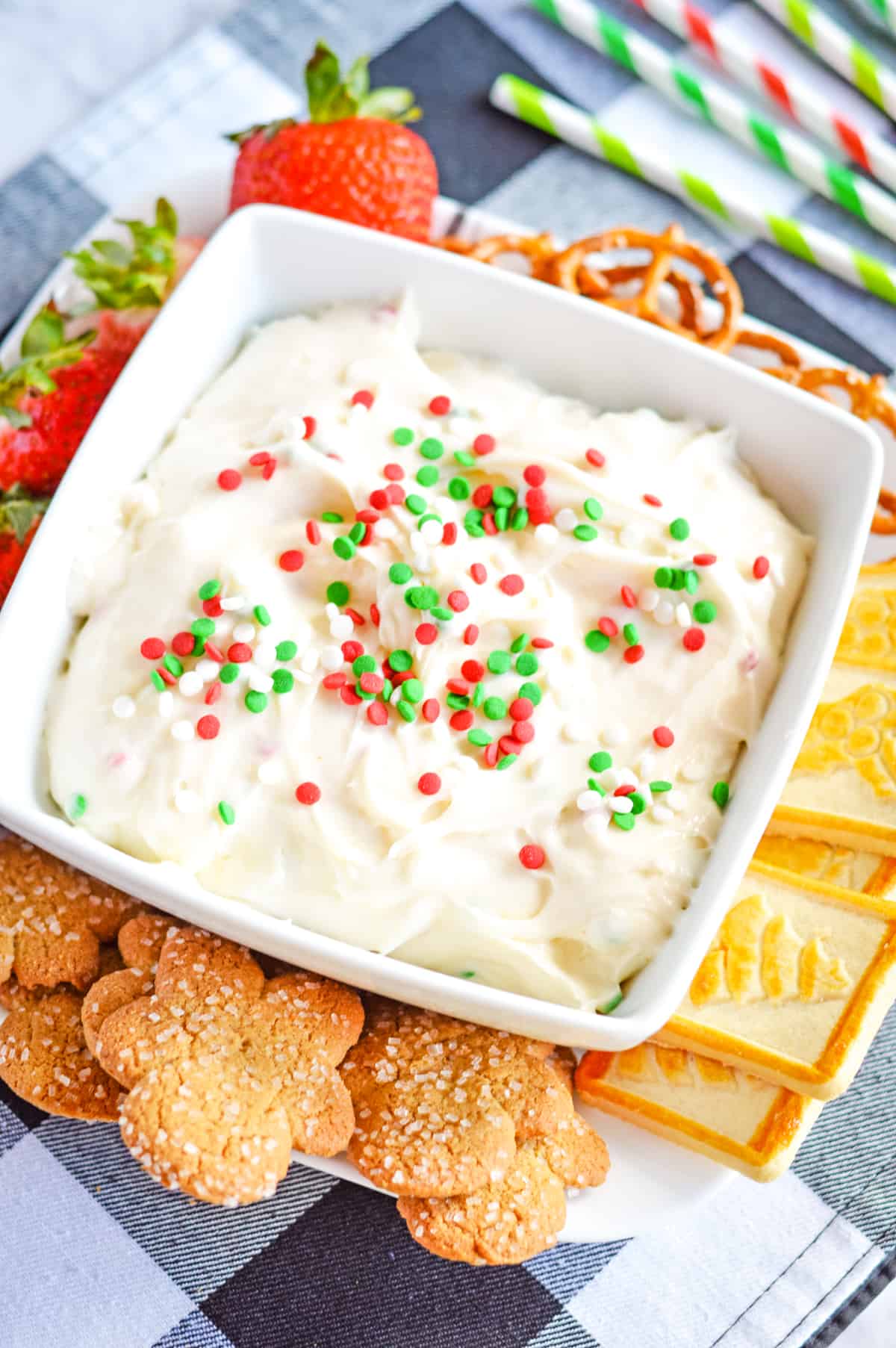 Christmas cookie dip in white bowl topped with Christmas sprinkles and served with Christmas cookies, pretzels, and strawberries for dipping.