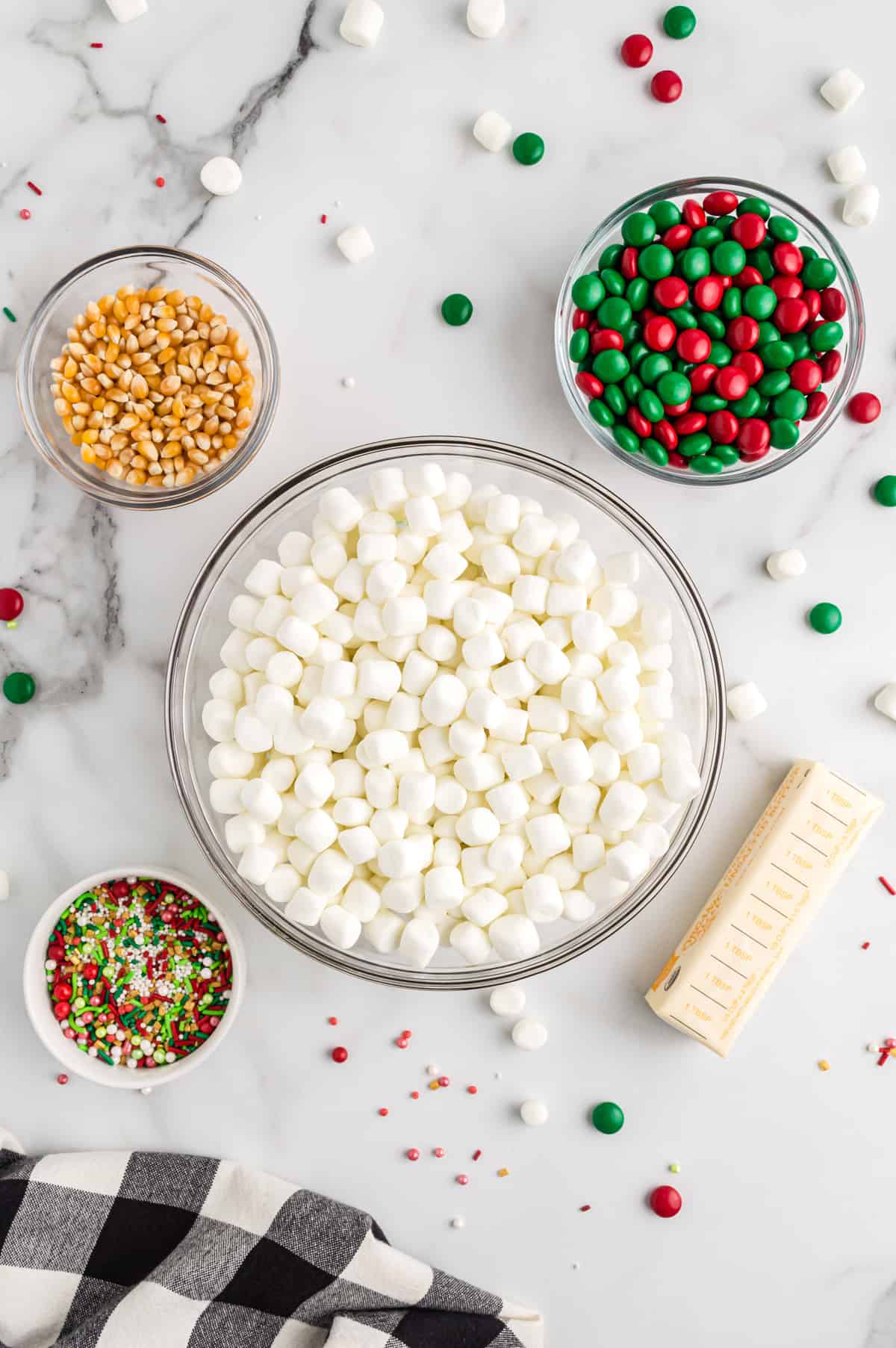 bowl of unpopped popcorn kernels, bowl of mini marshmallows, bowl of red and green chocolate candies, stick of butter, bowl of sptinkles
