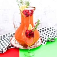 Christmas Punch with vodka and cranberry in a large pitcher garnished with cranberries, orange slice, and rosemary. A glass of the cranberry punch is in front of the pitcher and also garnished with cranberries, orange, and rosemary