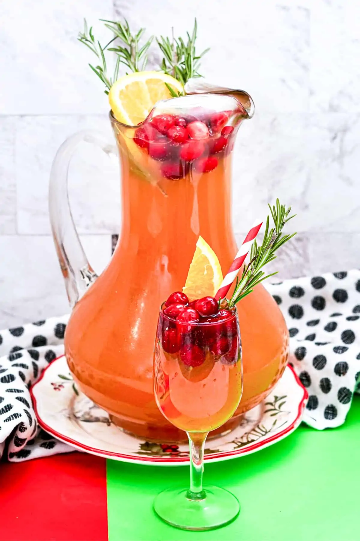 Christmas Punch with vodka and cranberry in a large pitcher garnished with cranberries, orange slice, and rosemary. A glass of the cranberry punch is in front of the pitcher and also garnished with cranberries, orange, and rosemary