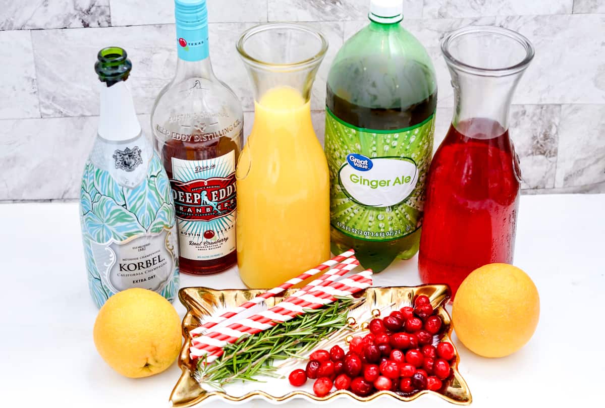 bottles of extra dry korbel champagne, cranberry vodka, orange juice, ginger ale, cranberry and juice on countertop. In front of the bottles are two oranges, a handful of fresh cranberries, a few sprigs of rosemary, and red and white striped paper straws