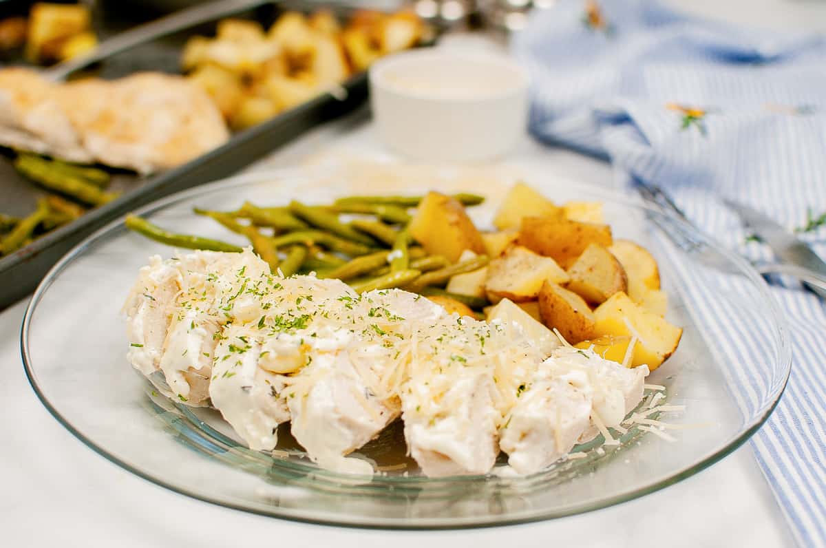 Creamy garlic herb chicken with green beans, and chopped potatoes on a clear plate with sheet pan with additional servings in background