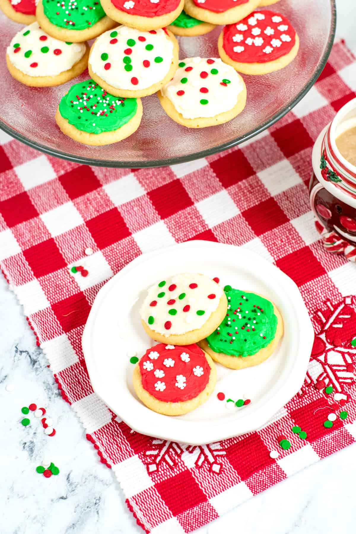 Soft Christmas Cookies with Frosting and sprinkles on white plate with platter of cookies and cup of coffee in background