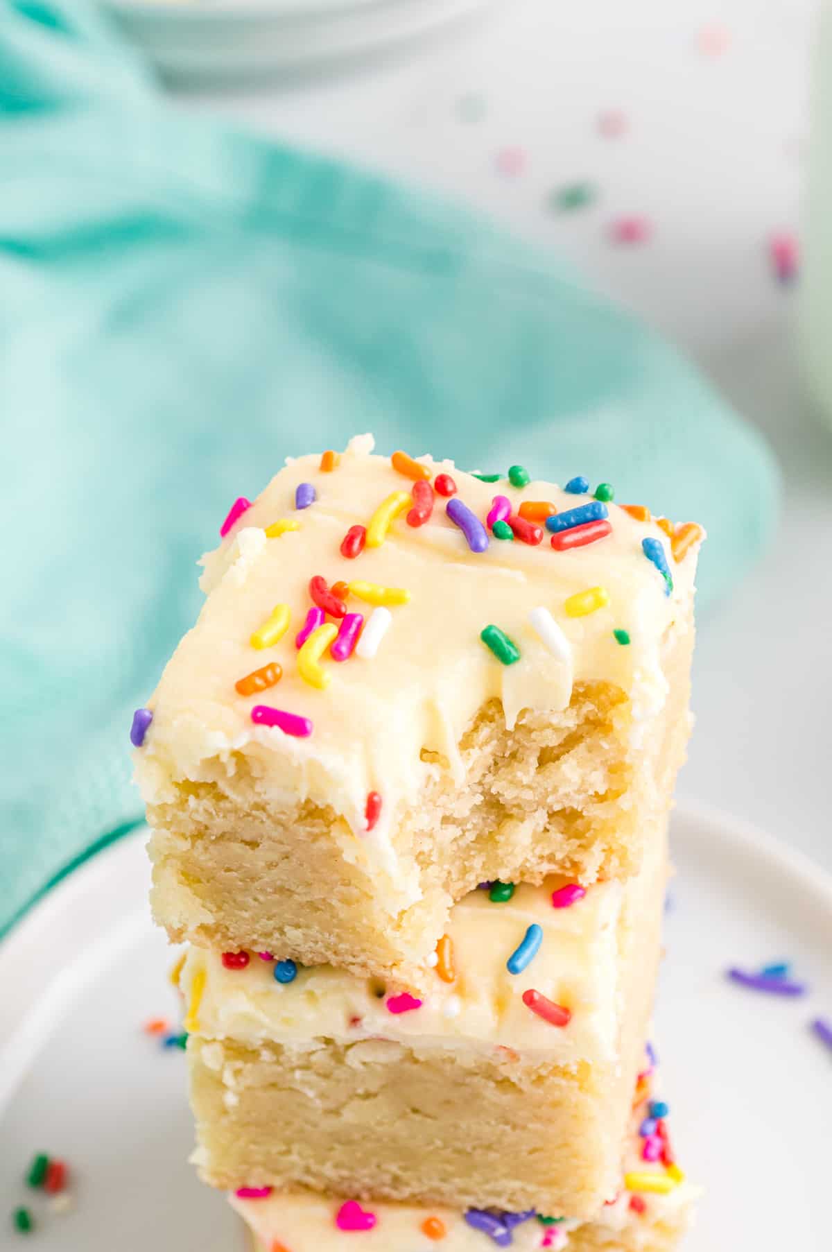 Frosted Sugar Cookie Bars with Sprinkles stacked on top of one another. The top bar has a bite taken out of it to show its thick and tender interior.