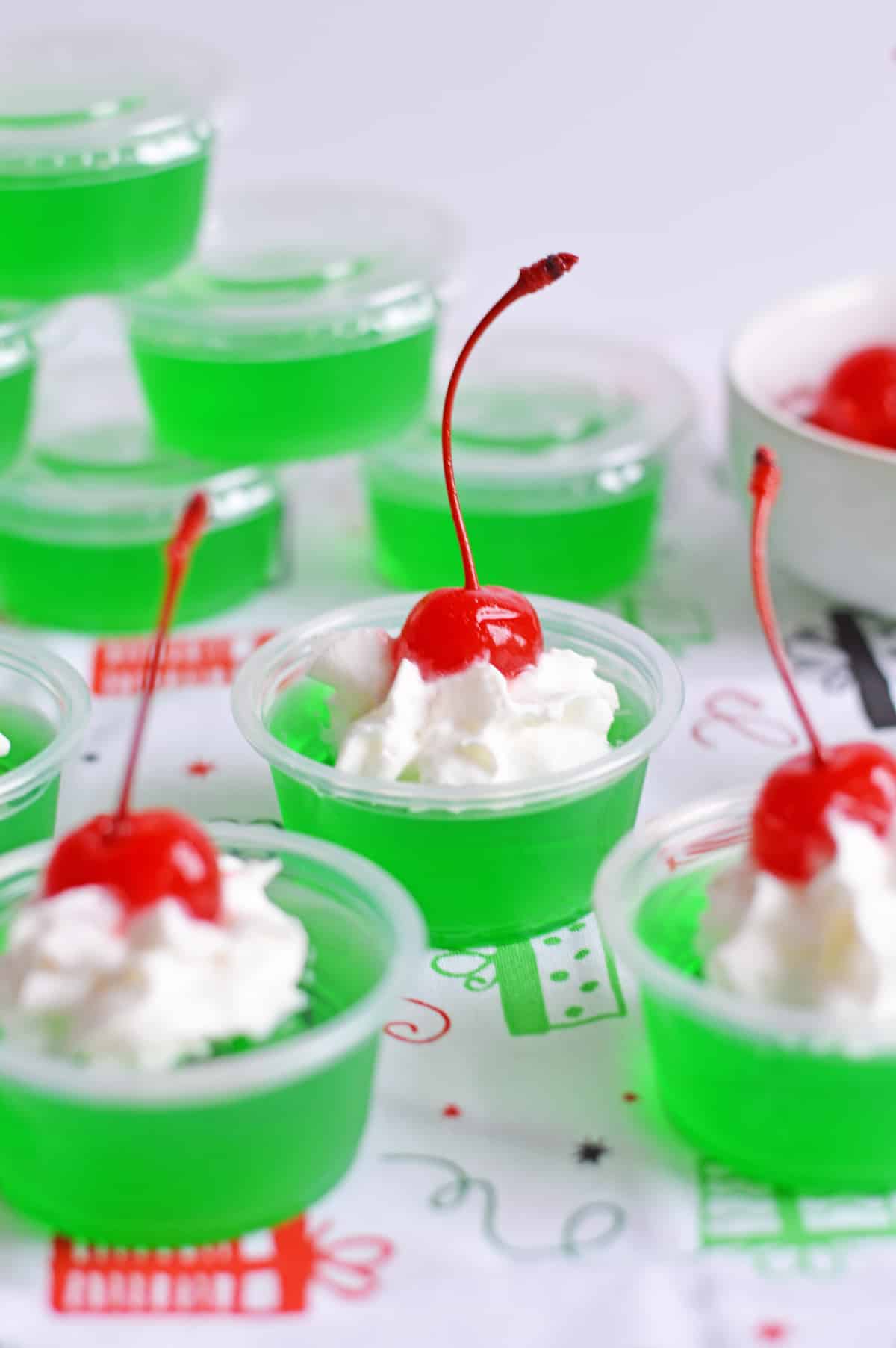 Grinch Jello Shots in small plastic shot cups made with lime jello, tequila, whipped cream, and a cherry on top