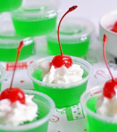 Grinch Jello Shots made with lime jello, tequila, and topped with whipped cream and a maraschino cherry.