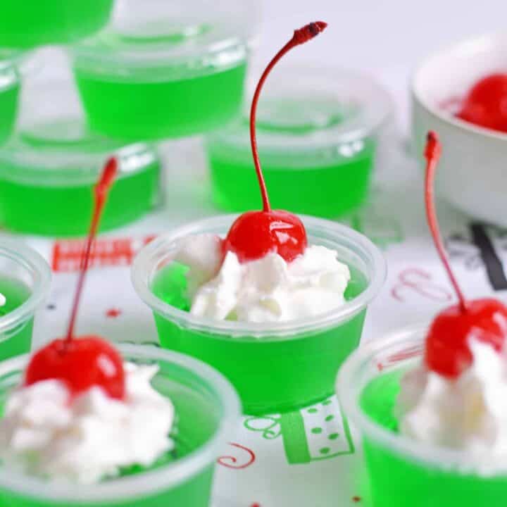 Grinch Jello Shots made with lime jello, tequila, and topped with whipped cream and a maraschino cherry.