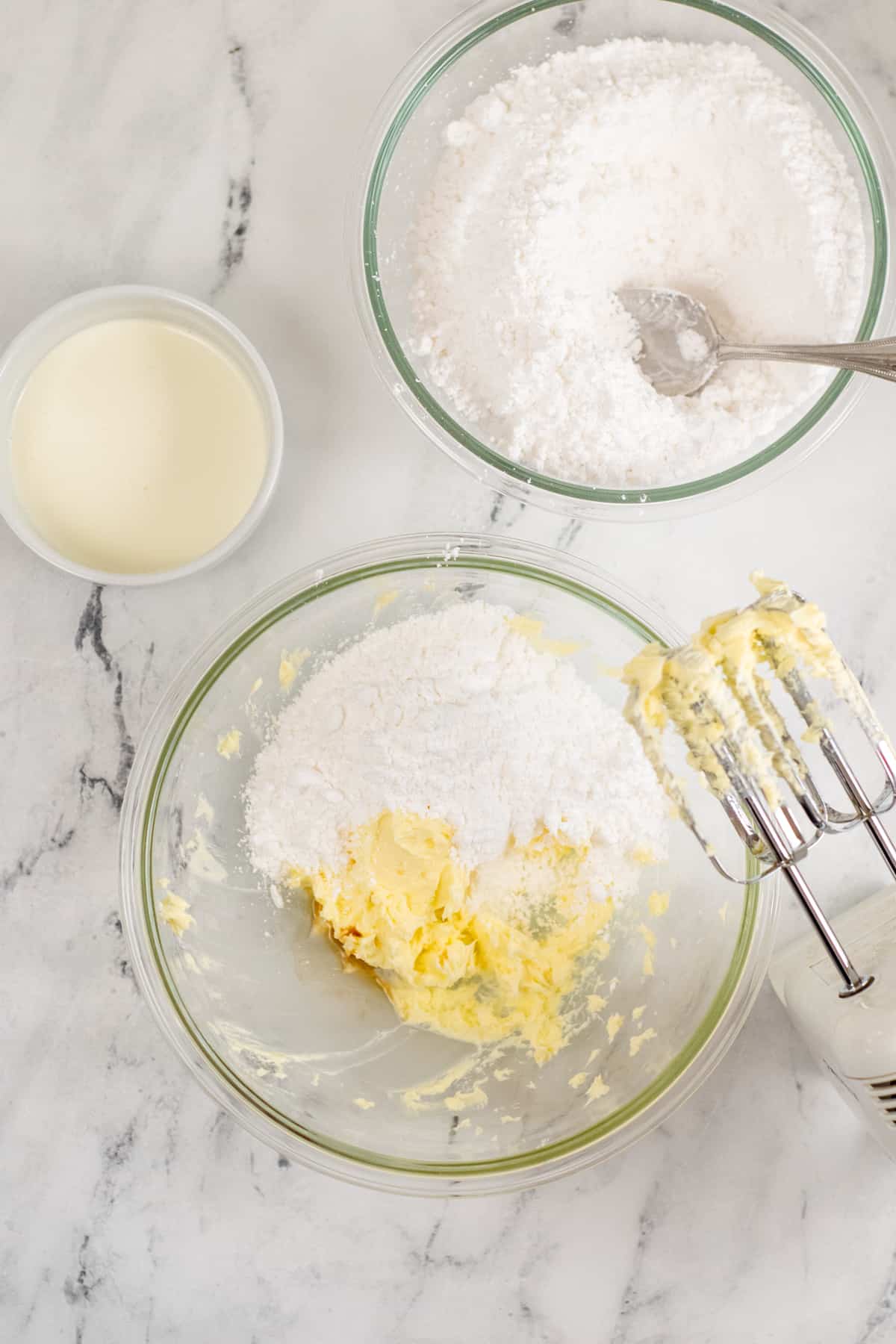 Large glass mixing bowl with creamed butter, powdered sugar, and hand mixer