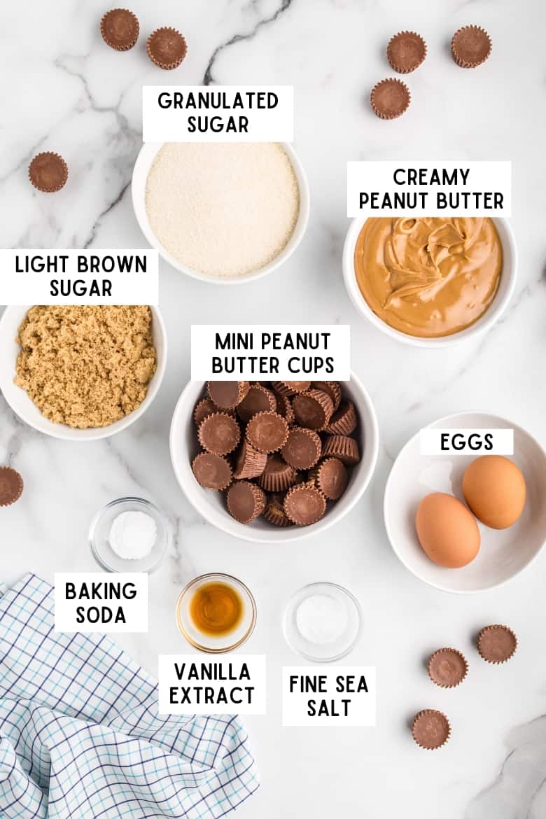 Ingredients on countertop: bowl of granulated sugar, bowl of creamy peanut butter, bowl of light brown sugar, bowl of unwrapped mini reese's peanut butter cups, 2 eggs, baking soda, vanilla, and fine sea salt. Several mini peanut butter cups and a kitchen towel surround the ingredients