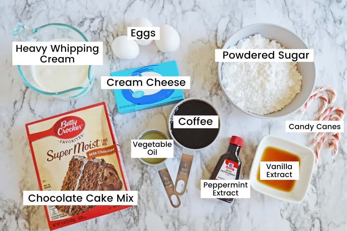 Ingredients on countertop: bowl of heavy whipping cream, 3 eggs, block of cream cheese, bowl of powdered sugar, candy canes, bowl of vanilla extract, bottle of peppermint extract, measuring cup with coffee, measuring cup with vegetable oil, and a box of Betty Crocker Milk Chocolate Super Moist Cake Mix