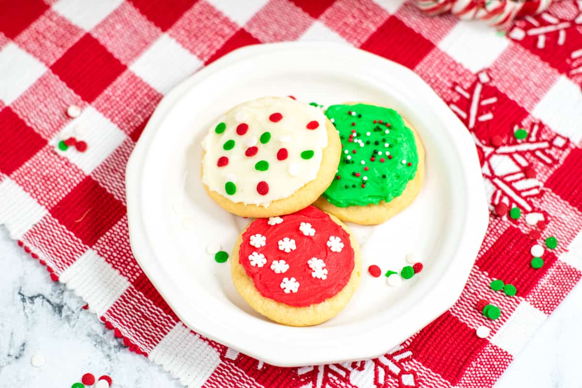 white dessert place with three frosted Christmas cookies, one red with white sprinkles; one green with green, white and red sprinkles; and one white with green and red confetti sprinkles.