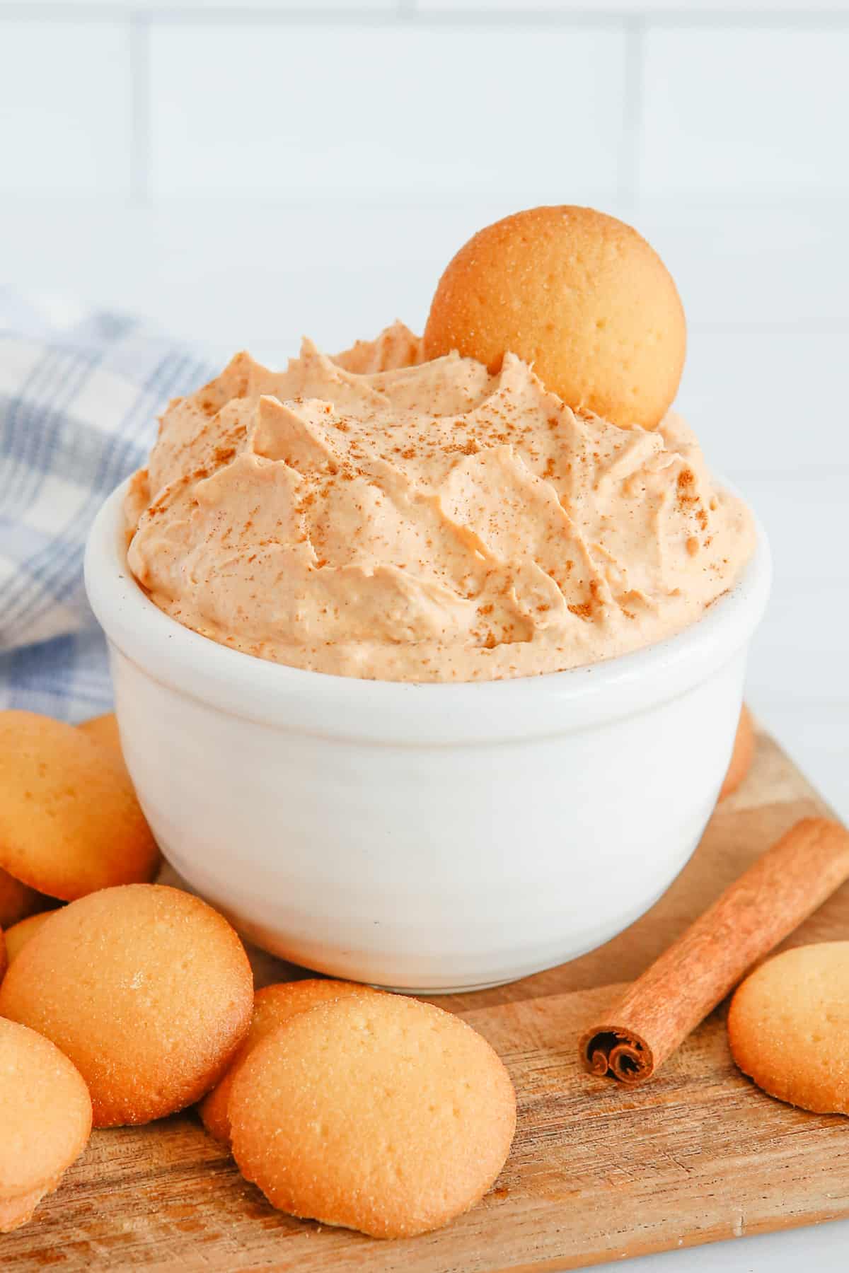 Pumpkin dip in white bowl with vanilla wafer sticking out of the top. Vanilla cookies and cinnamon sticks are on tabletop around the bowl.