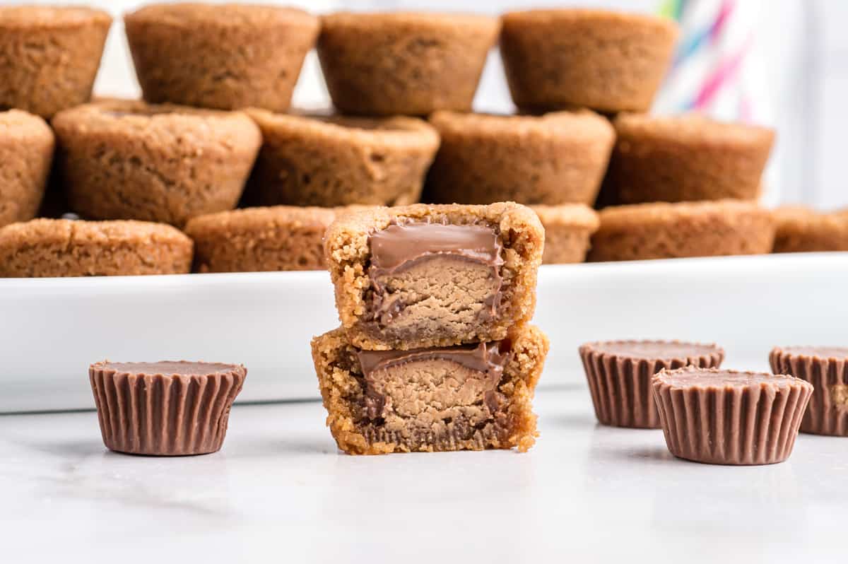 peanut butter cookies with peanut butter cups pressed in center. Several cookies are stacked neatly on platter and one is removed and cut in half to show inside. 