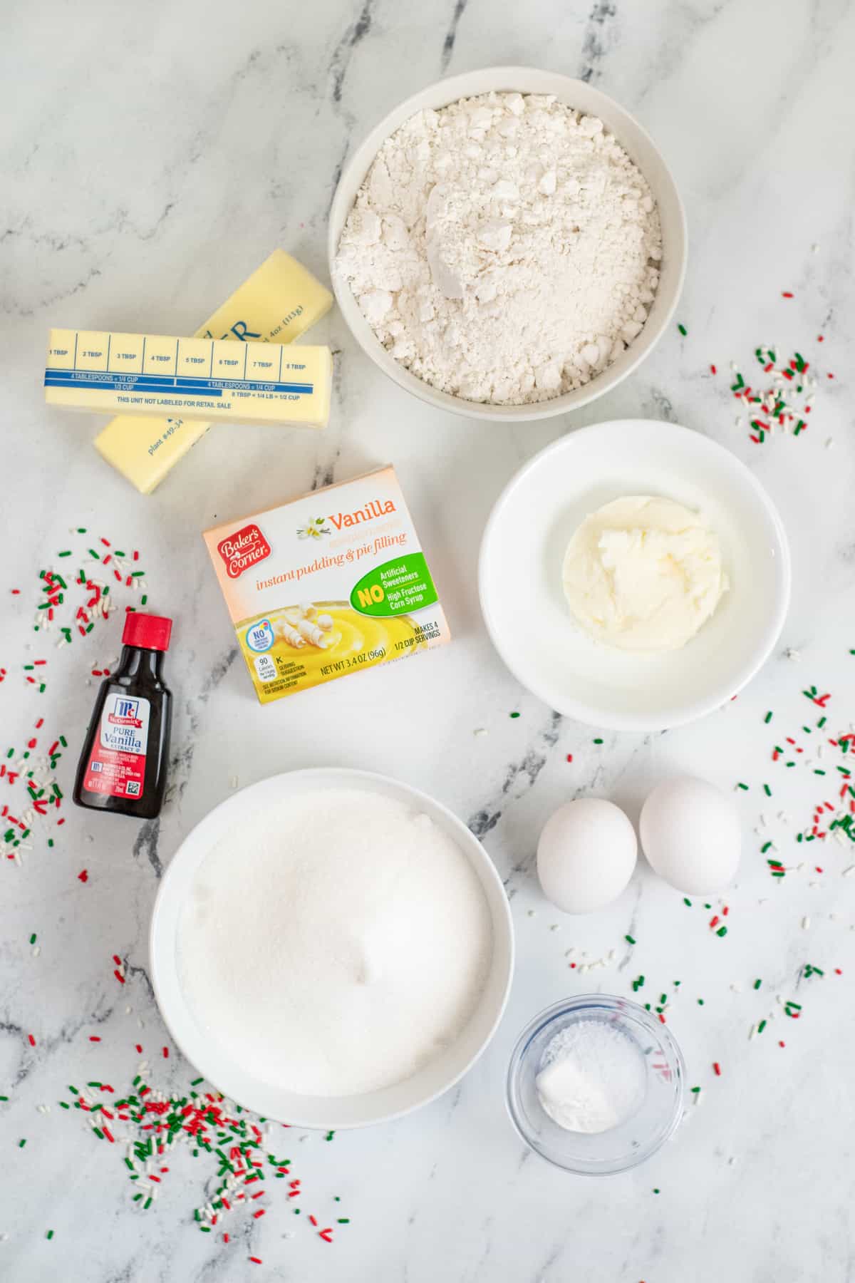 Two sticks of butter, bowl of flour, bowl of shortening, 2 eggs, box of vanilla pudding mix, bowl of granulated sugar, baking powder, bottle of vanilla, and red, white, and green sprinkles scatted all around