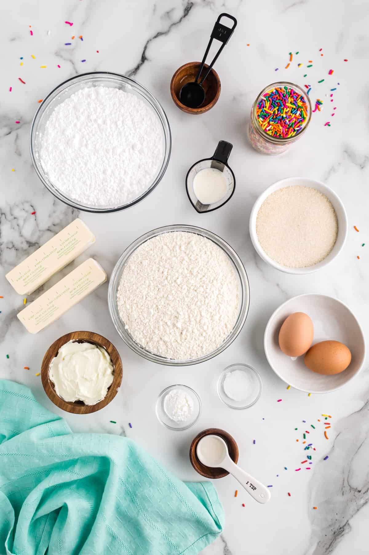 Ingredients on countertop: flour, white sugar, 2 sticks of butter, shortening, clear vanilla extract, almond extract, 2 eggs, salt, making powder, powdered sugar, cream, sprinkles