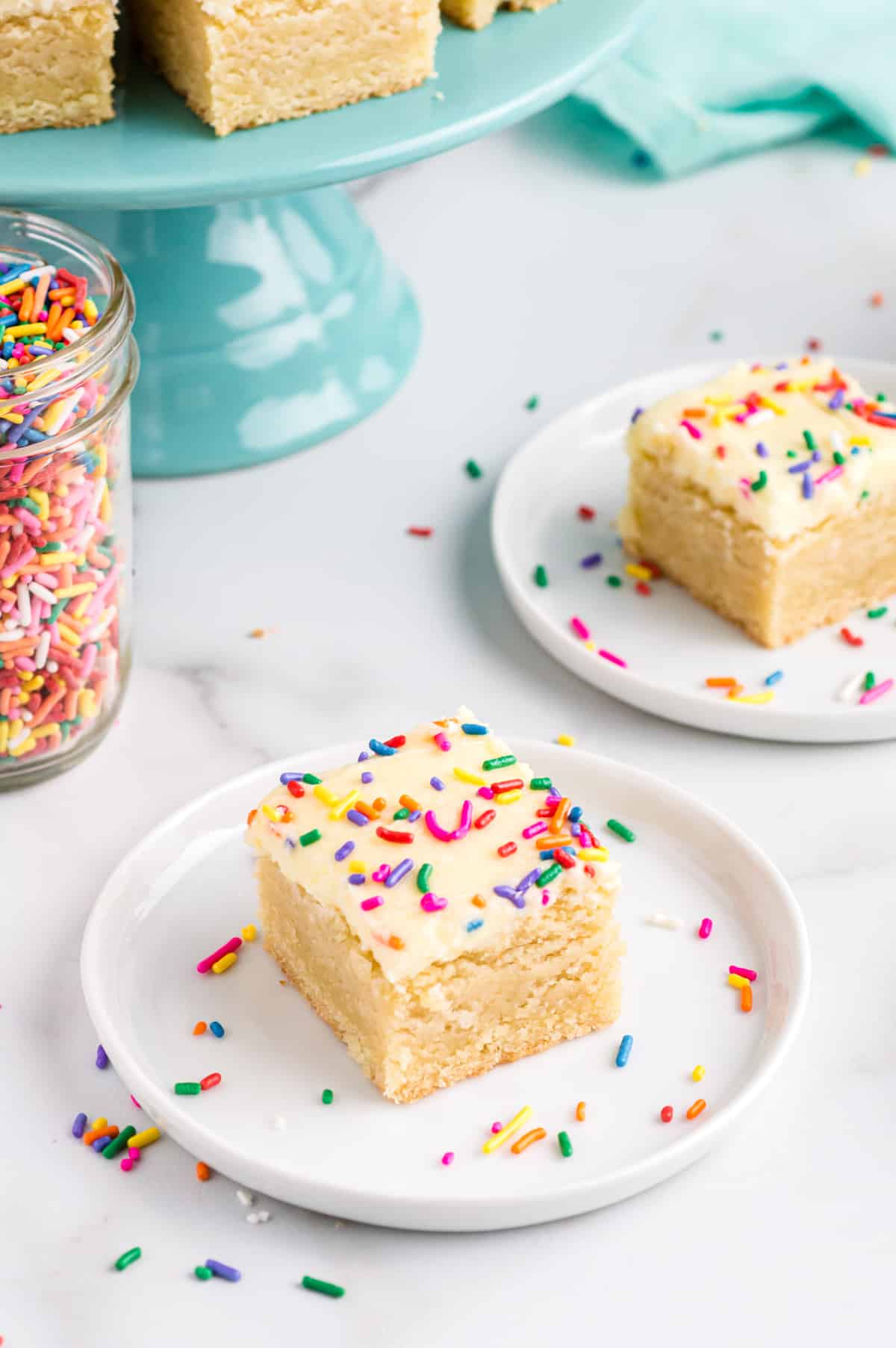 Two sugar cookie bars with frosting and sprinkles on white plates. Jar of rainbow sprinkles and more bars are in background.