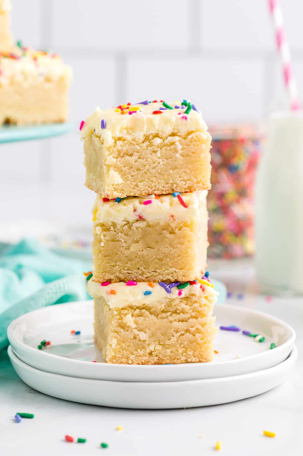 3 thick sugar cookie bars with frosting stacked on a white plate. A jar of rainbow sprinkles can be seen in background.