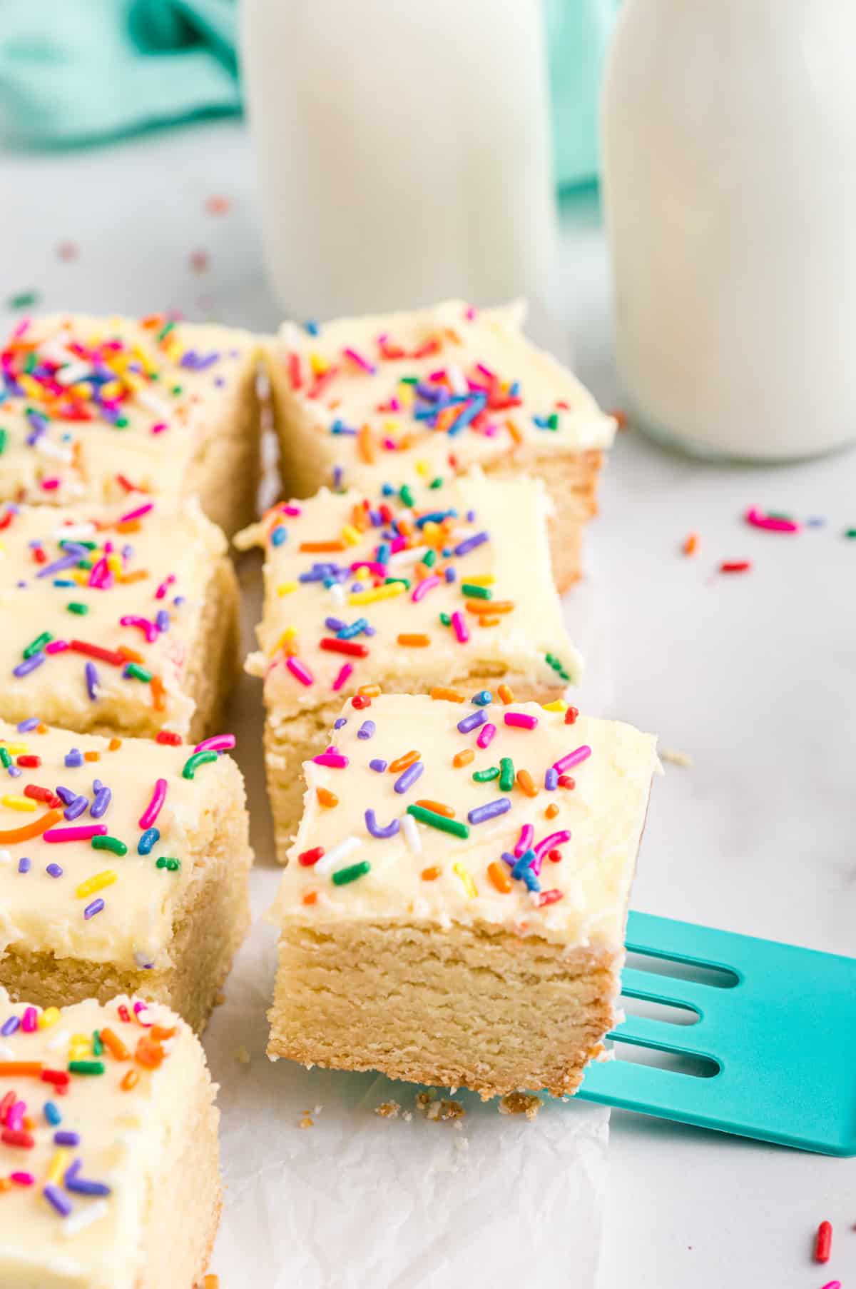 Sugar Cookies Bars with vanilla frosting and rainbow sprinkles, cut into squares and being served with a teal spatula. Milk in glasses can be seen behind the bars.