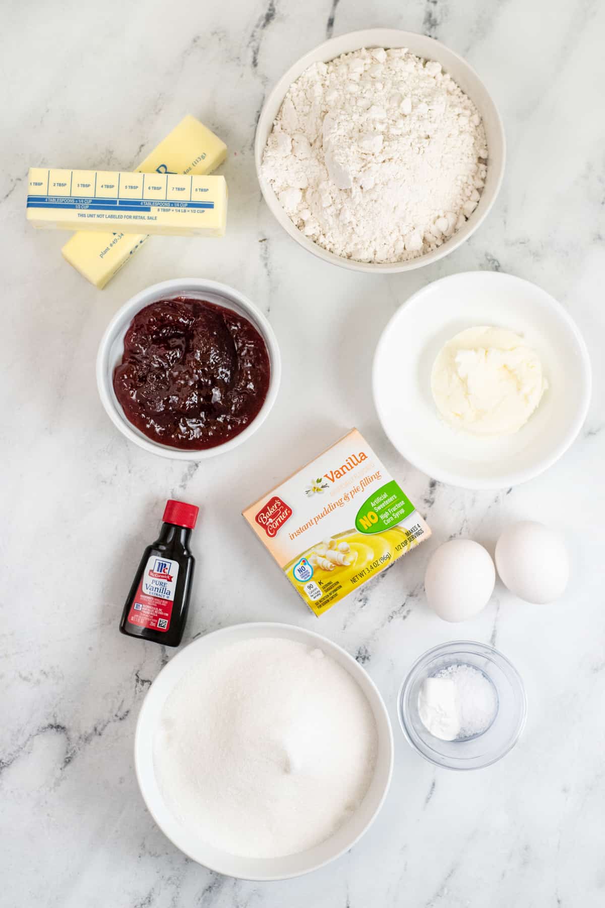 Ingredients on countertop: two sticks of butter, bowl of flour, bowl of shortening, 2 eggs, box of vanilla pudding mix, salt, baking powder, granulated sugar, bowl of strawberry jam