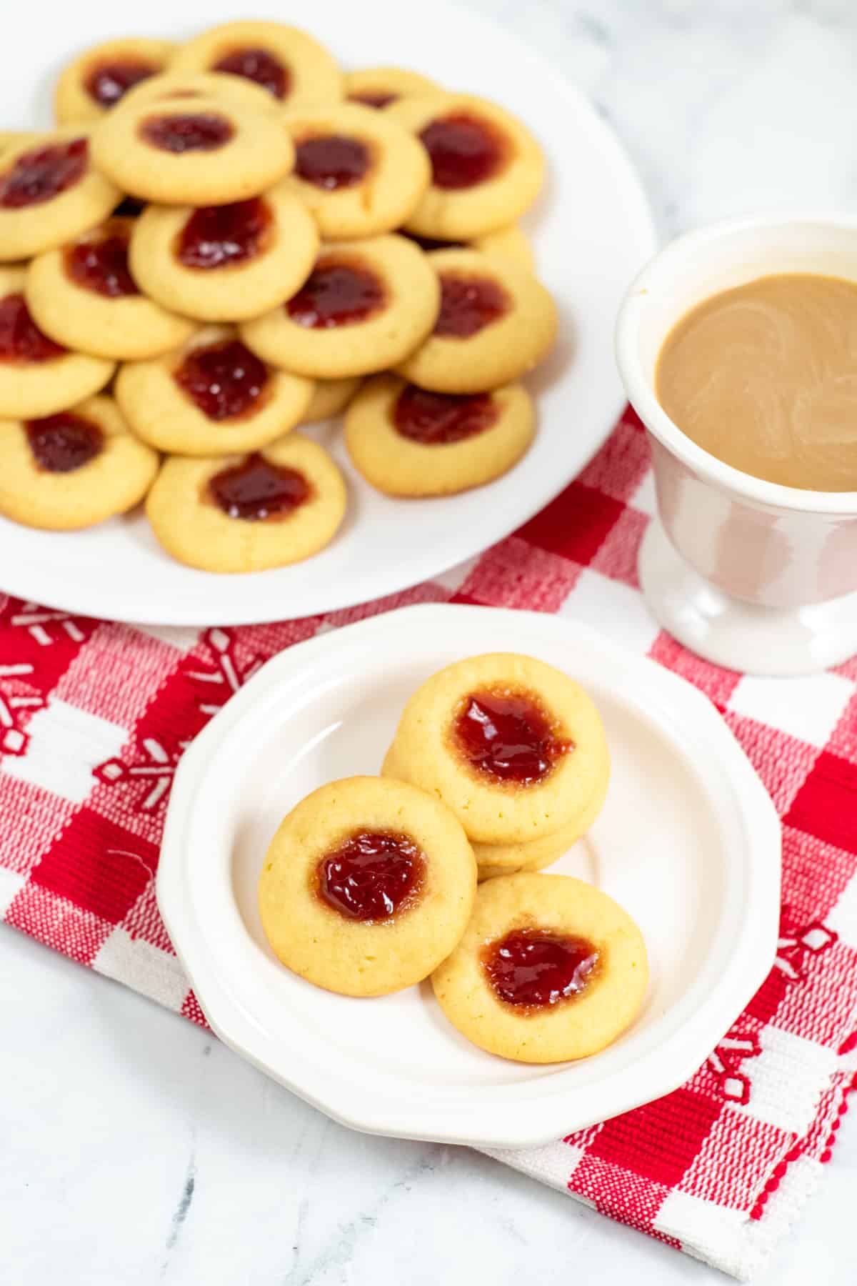 white plate with four thumbprint cookies and a mug of coffee and larger plate with thumbprint jam cookies piled high in background