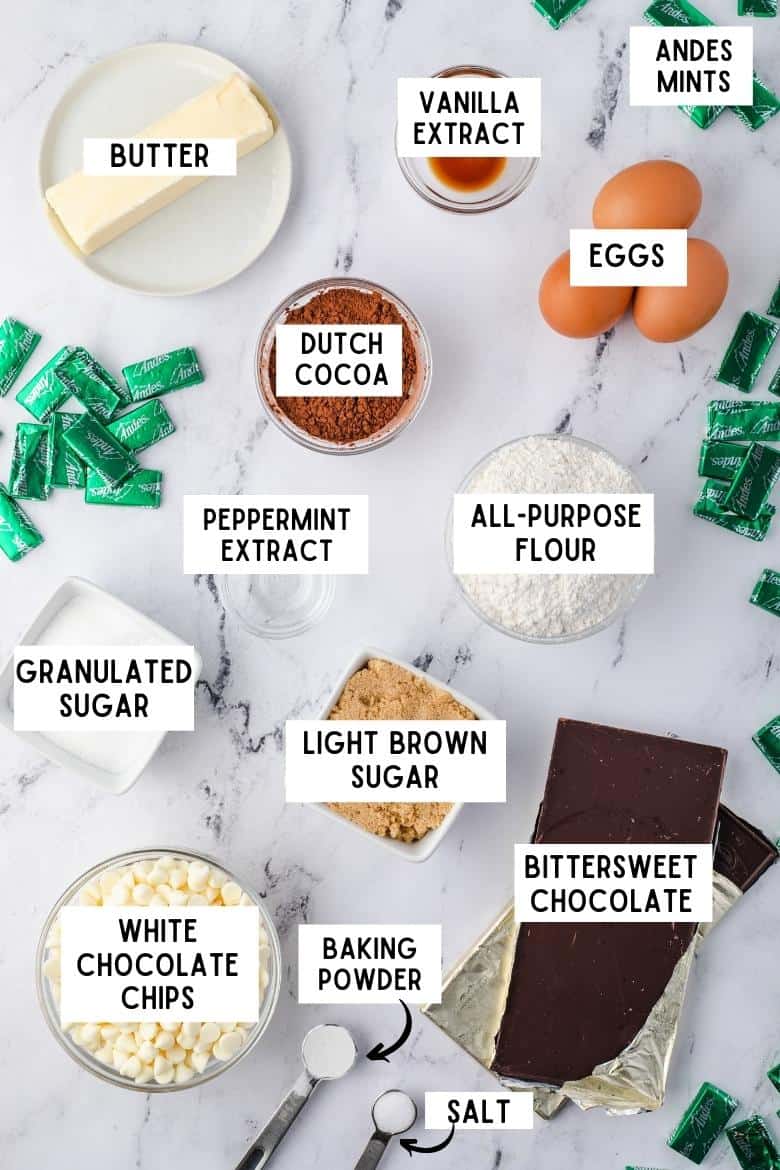 Ingredients on countertop: stick of butter, vanilla extract, andes mints, dutch cocoa powder, 3 eggs, all-purpose flour, salt, baking powder, granulated sugar, light brown sugar, peppermint extract, white chocolate, and bittersweet chocolate bars