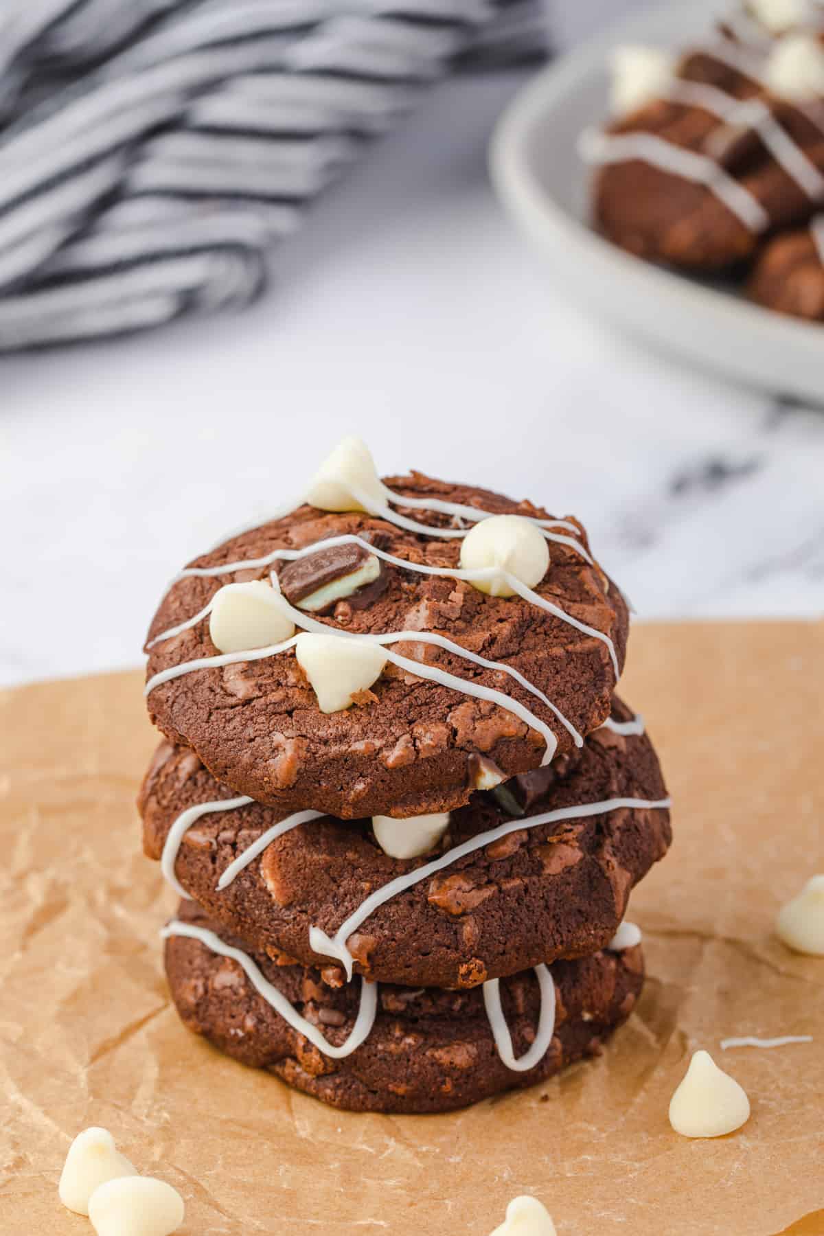 Three stacked mint chocolate brownie cookies with white chocolate chips, chunks of andes mints, and white chocolate drizzle.